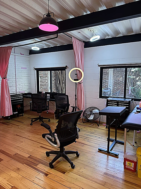 Sala de tatuaje con lámparas colgantes rosas, cortinas rosas, muros blancos y piso de madera.