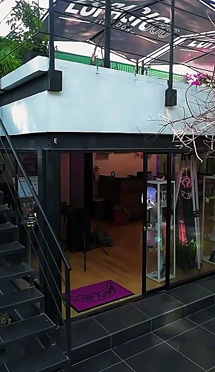 Fachada del estudio: estructura de acero negro con escalera y terraza arriba, rodeada de plantas.
