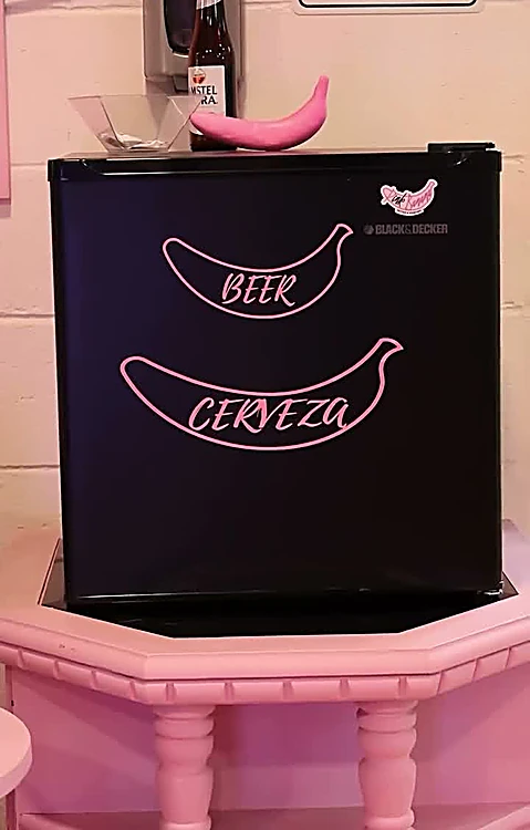 Frigobar negro con plátanos dibujados en rosa y las palabras Beer y Cerveza, sobre un mueble rosa.