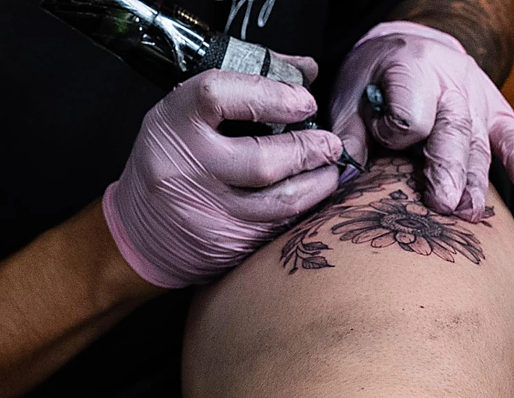 Tatuador con guantes rosas trabajando flores en sombra sobre el muslo.