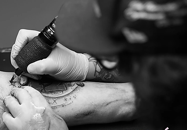 Manos con guantes y la máquina de tatuar trabajando sobre un brazo, foto en blanco y negro.
